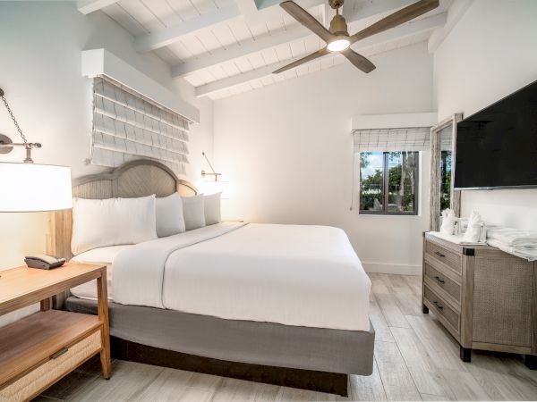 A modern bedroom with a large bed, bedside tables, a ceiling fan, a wall-mounted TV, and a window overlooking greenery.