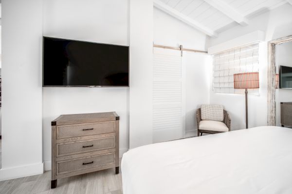 A minimalist bedroom featuring a mounted TV, a wooden dresser, a cozy chair, a floor lamp, and a bed with white linens.