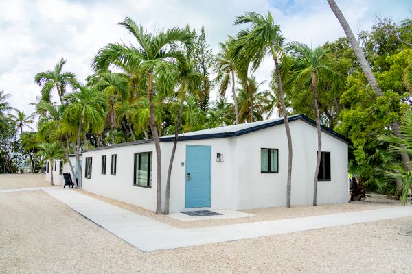 A modern white house with a blue door, surrounded by palm trees, on a gravel pathway under a partly cloudy sky.