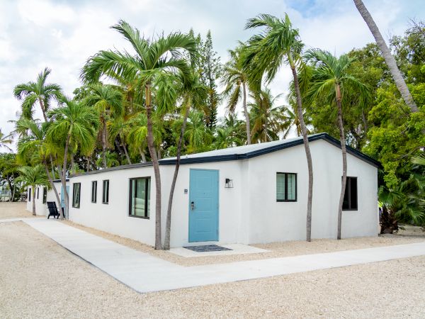 A modern white house with a blue door, surrounded by palm trees, on a gravel pathway under a partly cloudy sky.