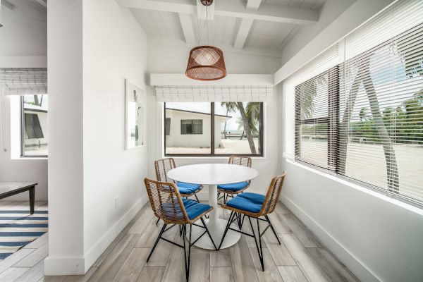 A bright dining area with a round table, four chairs, large windows, blue cushions, and a view of palm trees outside completes the scene.