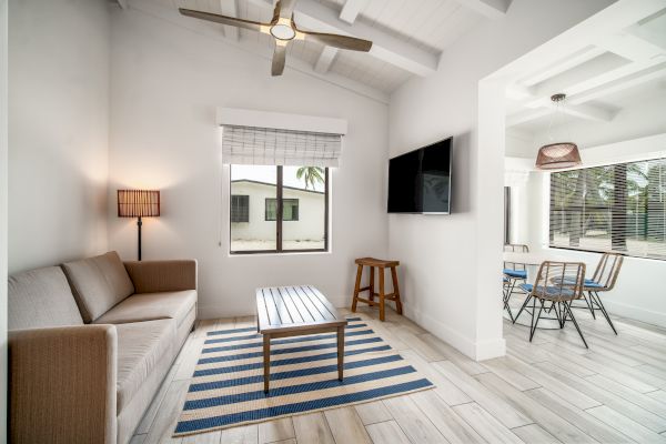 A modern living room with a beige sofa, striped rug, and TV. Dining area with table and chairs on the right. Ceiling fan and large windows.