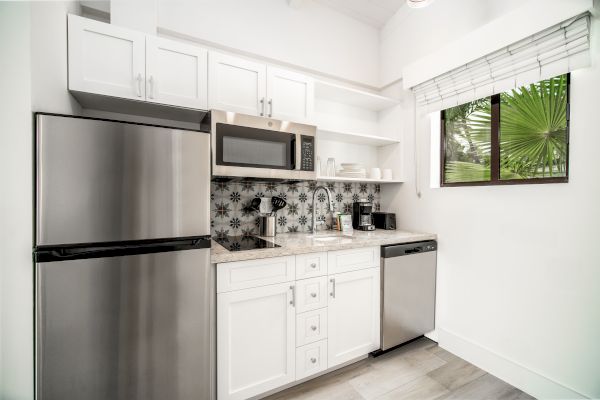 A modern kitchen with a stainless steel fridge, microwave, and dishwasher. White cabinets, a patterned backsplash, sink, and a window are present.