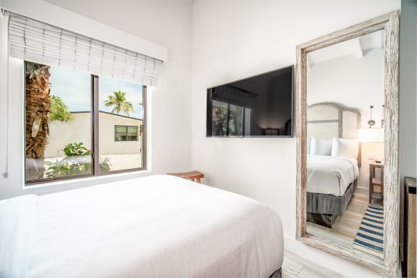 A modern bedroom with a white bed, large window, wall-mounted TV, and full-length mirror reflecting another bed and nightstand.