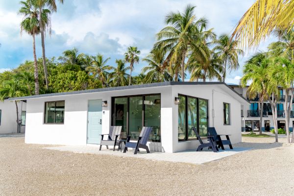 A small white house with large windows, surrounded by palm trees, featuring outdoor seating on a gravel yard under a clear blue sky.