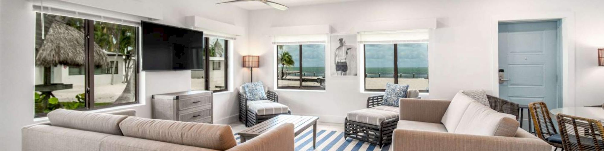 A cozy living room features two sofas, a striped rug, large windows, and a view of the ocean. There's a ceiling fan and modern decor.