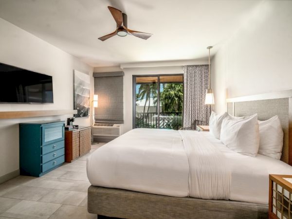 A modern hotel room with a large bed, TV, ceiling fan, and a balcony view with trees outside; simple and well-lit design.