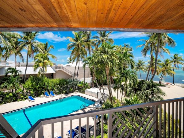 A tropical view of a pool surrounded by palm trees, blue lounge chairs, and beachfront with ocean in the background under a sunny sky.