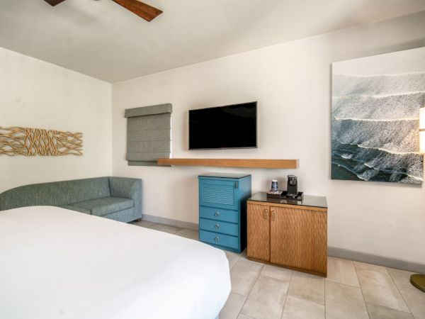 The image shows a modern hotel room with a bed, a wall-mounted TV, a blue chest, a gray sofa, abstract artwork, and a floor lamp.