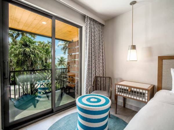 A bright hotel room features a bed, side table, and a striped ottoman. A balcony overlooks palm trees through glass doors.
