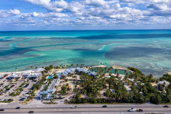 A coastal area with turquoise waters, white buildings, palm trees, and a parking lot, viewed from above.