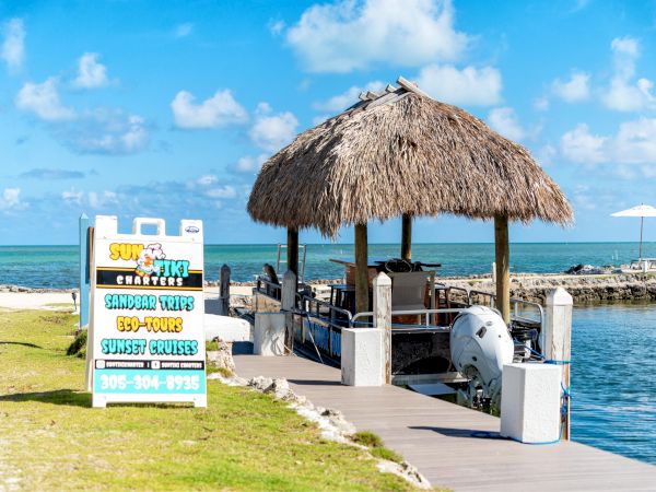 A thatched hut with a boat dock, near a sign for water activities like tours and cruises, set by the ocean under a bright blue sky.
