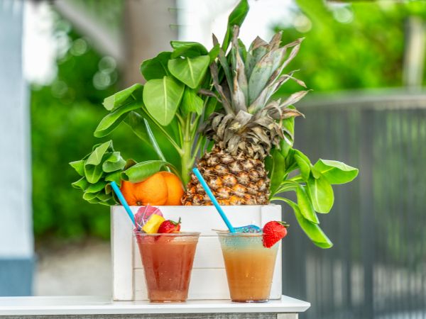 Two tropical drinks with straws, garnished with fruit, are next to a box containing a pineapple, oranges, and leafy greens.