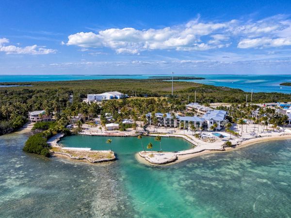 Aerial view of a coastal resort with palm trees, surrounding water, and distant land under a blue sky with scattered clouds.