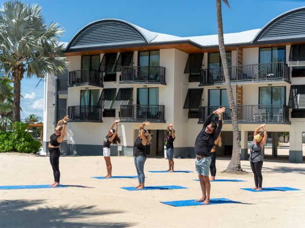 People are practicing yoga outdoors on blue mats in front of a modern building with palm trees around, under a clear blue sky.