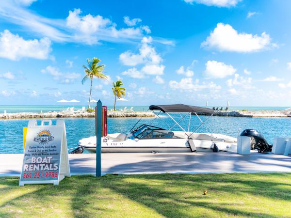 A sunny waterfront scene with a moored boat and a sign advertising boat rentals, set in a tropical locale with blue skies and palm trees.