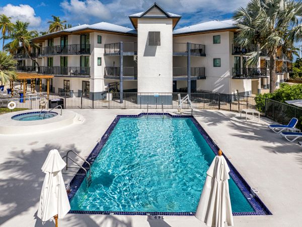 A swimming pool beside an apartment building, with lounge chairs, umbrellas, a hot tub, and palm trees under a blue sky.