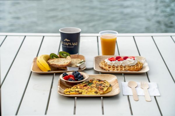 A breakfast spread with an omelet, waffle, fruits, juice, and coffee on a table near water, along with wooden cutlery arranged neatly.