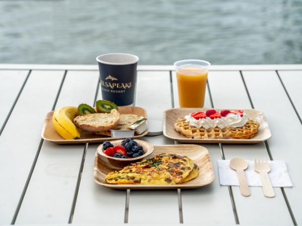 A breakfast spread with an omelet, waffle, fruits, juice, and coffee on a table near water, along with wooden cutlery arranged neatly.
