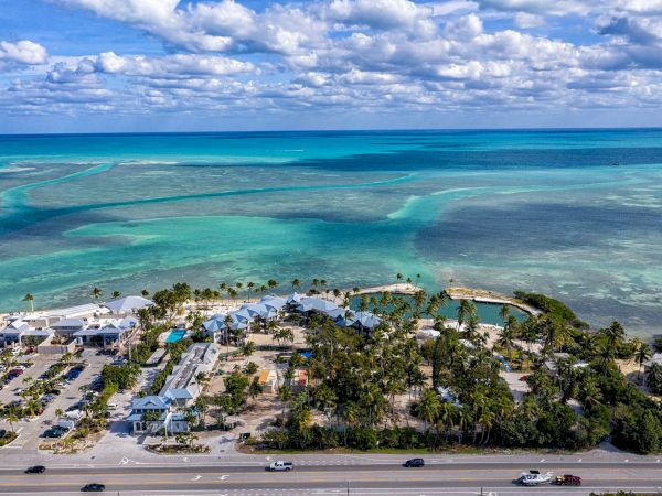 Aerial view of a tropical beachfront resort with palm trees, turquoise shallows, and a cloudy blue sky over the ocean.