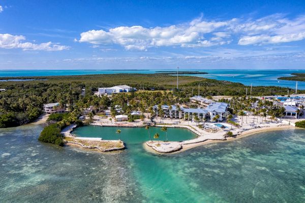 Aerial view of a coastal resort with a lagoon, palm trees, and buildings surrounded by turquoise water and lush greenery under a blue sky.
