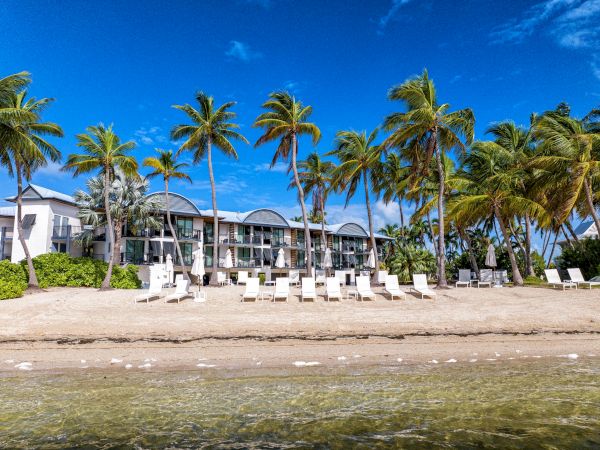 A beachfront scene with palm trees, lounge chairs, and a building under a clear blue sky, viewed from the water’s edge.
