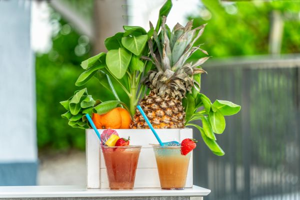 Two tropical drinks with straws sit in front of a crate containing a pineapple, orange, and green leaves, against a blurred background.