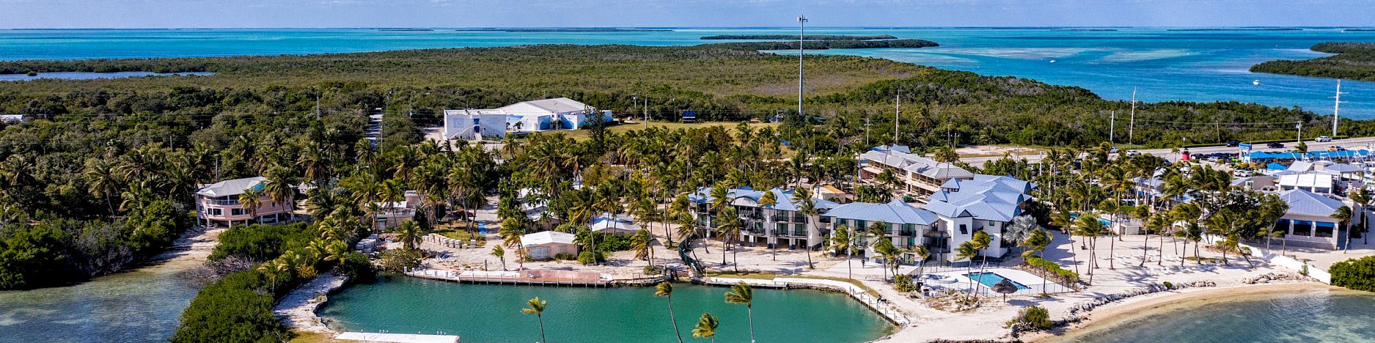 Aerial view of a seaside resort with clear turquoise water, white sandy beaches, and lush greenery under a blue sky with scattered clouds.