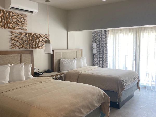 Bright modern hotel bedroom with two beds, ceiling fan/light, wall art, and large windows with patterned curtains.