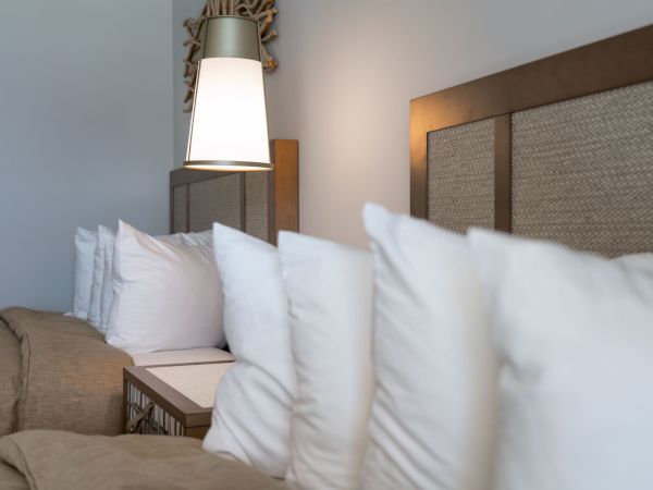 A cozy hotel bed with fluffy white pillows and a warm bedside lamp, framed by wooden headboard details.
