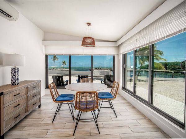 A bright patio dining area with a round table, rattan chairs, and ocean-and-pool views through large sliding windows.