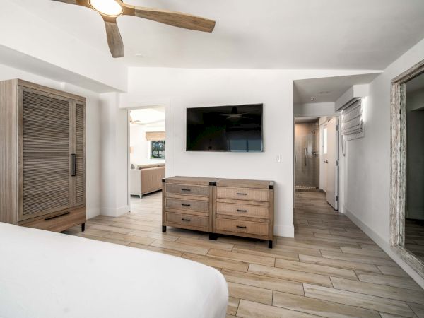 Bright modern bedroom with a wall-mounted TV, wood dresser, ceiling fan light, and open door to a bathroom.