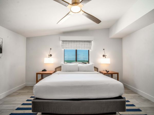 A bright bedroom with a large bed, ceiling fan, bedside lamps, and a window with blinds facing the ocean.