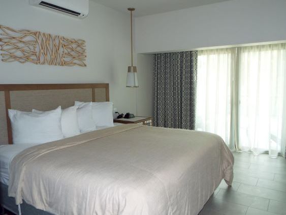 A tidy hotel bedroom with a large bed, air conditioner, bedside lamp, and bright windows with curtains.