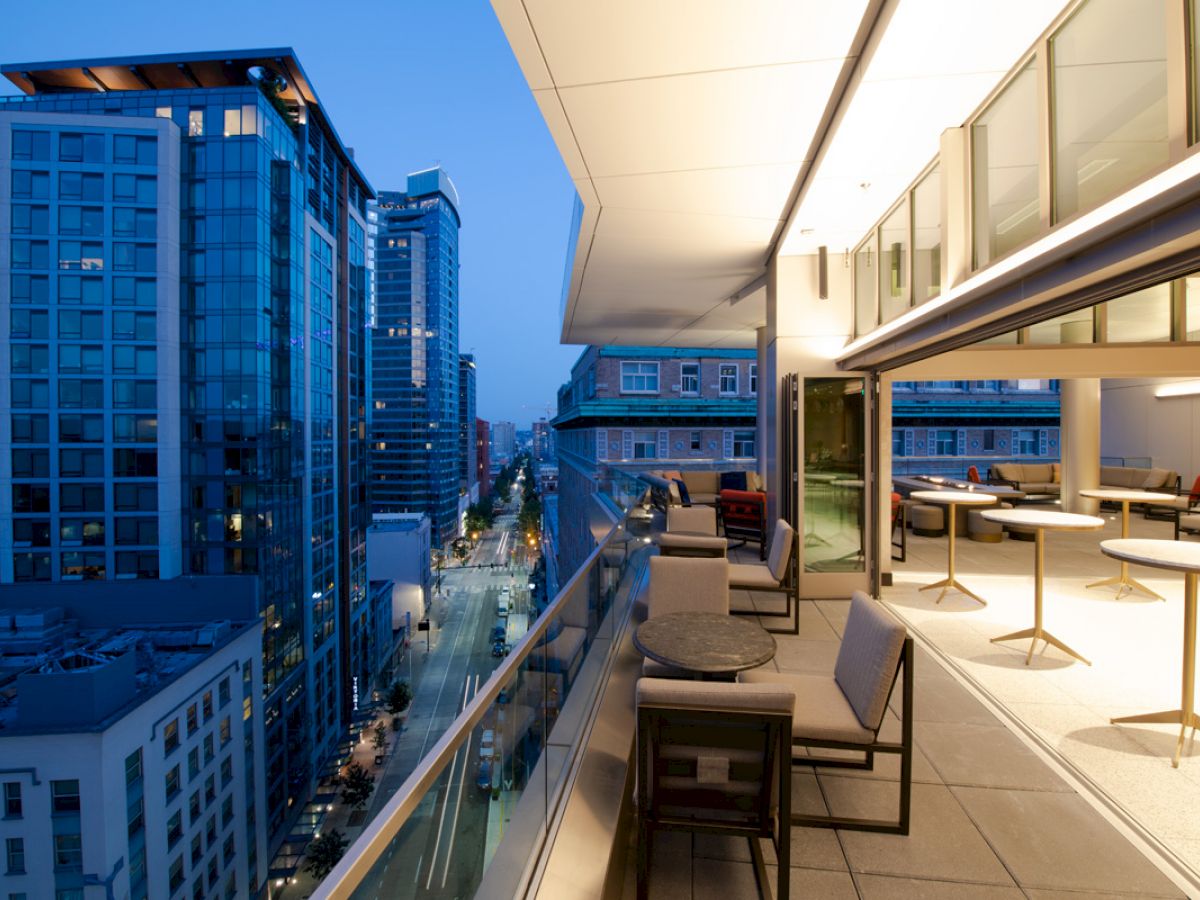 A modern outdoor balcony view with tables and chairs overlooking a cityscape of high-rise buildings during the evening.