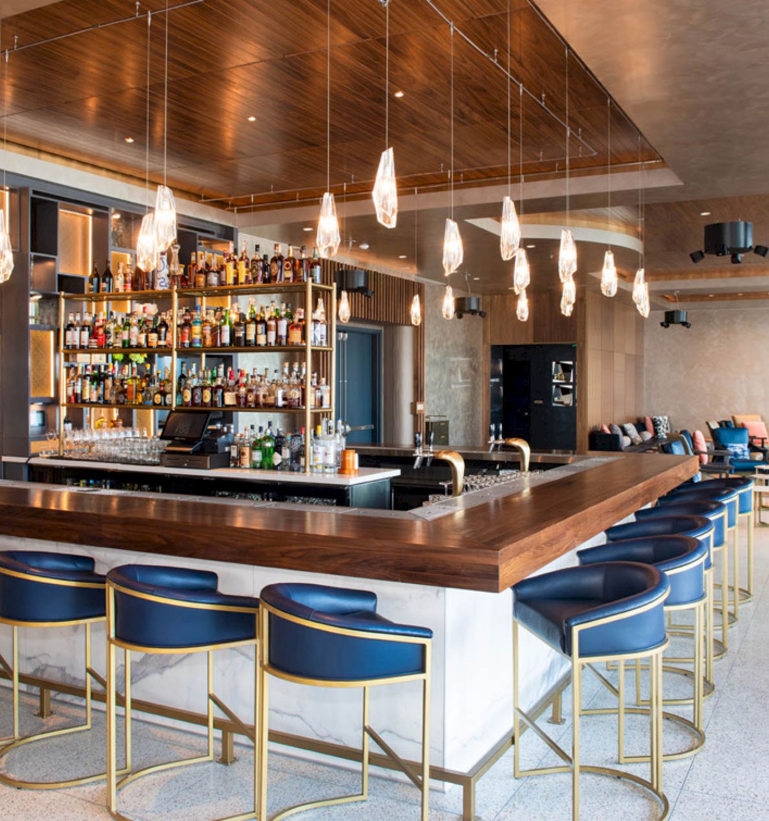 The image depicts a modern bar with a wooden counter, blue-gold stools, hanging lights, a stocked bar shelf, and a seating area in the background.