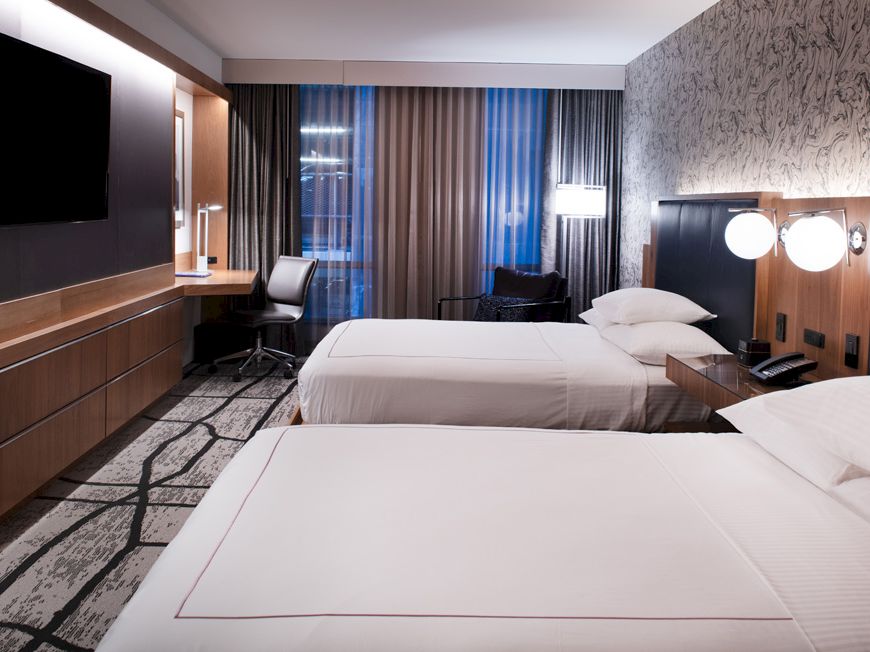 A modern hotel room with two neatly made beds, a wall-mounted TV, a desk with a chair, and large windows with curtains for natural light.