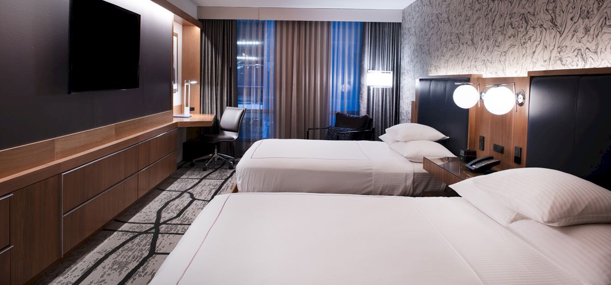 A modern hotel room with two neatly made beds, a wall-mounted TV, a desk with a chair, and large windows with curtains for natural light.