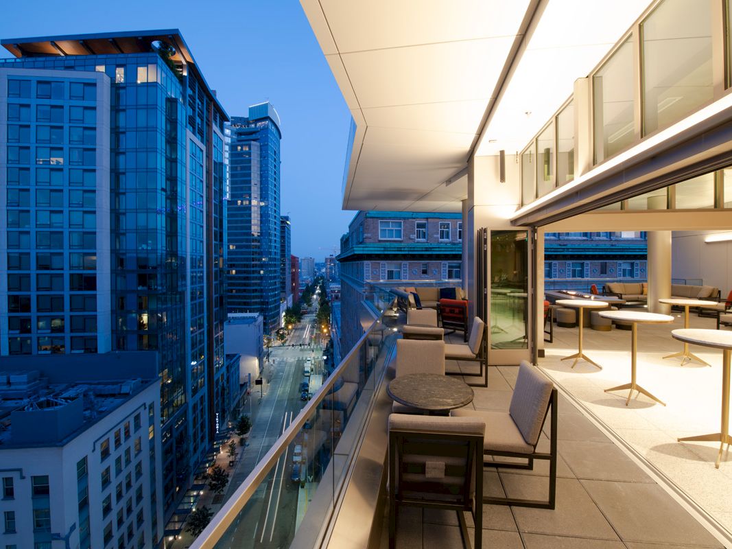 A modern balcony overlooking a cityscape at dusk, featuring patio furniture and an open-plan indoor space with more seating areas.