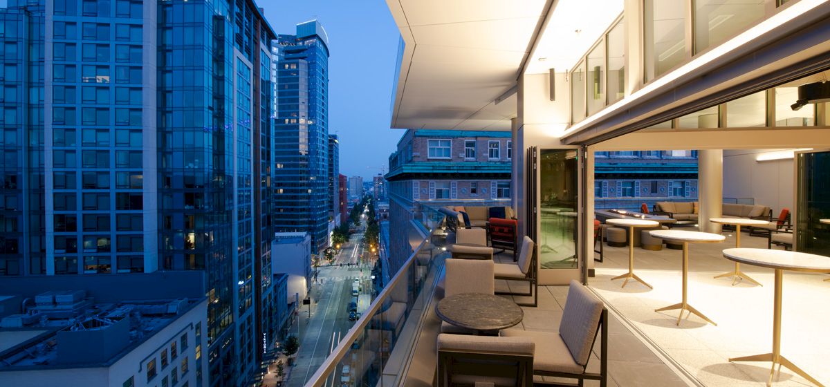 A modern balcony overlooking a cityscape at dusk, featuring patio furniture and an open-plan indoor space with more seating areas.