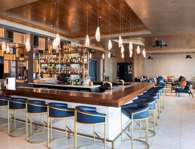 A modern bar with blue cushioned stools, hanging lights, and a well-stocked liquor display. The area includes some seating and contemporary decor.