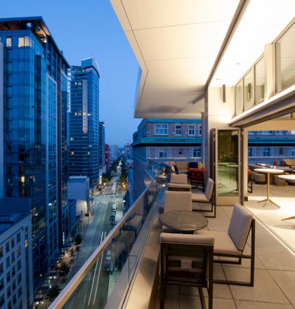 A modern urban balcony at twilight, overlooking a city street with tall buildings, featuring tables, chairs, and a glass railing.