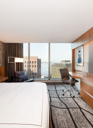 A modern hotel room with a king-size bed, large TV, desk, chair, and large window with a city view.