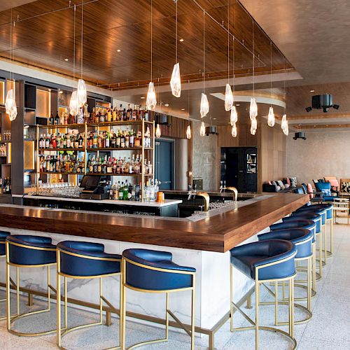 A modern bar with a wooden counter, blue bar stools, pendant lights, and a well-stocked shelf of liquor bottles, with seating areas in the background.