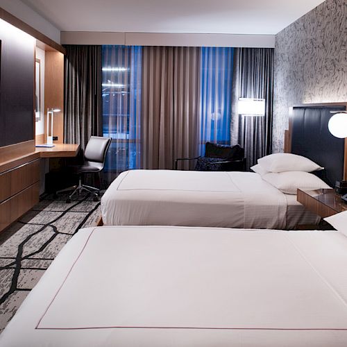 This image shows a modern hotel room with two beds, a flat-screen TV, a desk with a chair, and large windows with curtains.