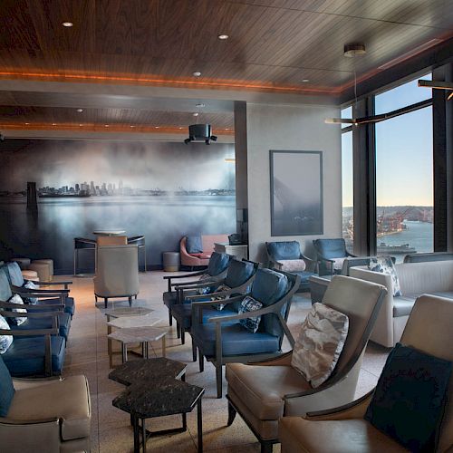 A modern lounge with comfortable seating, a large mural, and floor-to-ceiling windows offering a scenic waterfront view with a Ferris wheel in the distance.