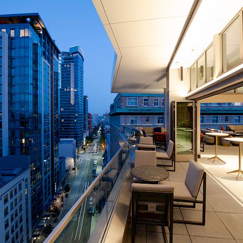 The image shows a modern rooftop terrace with seating and tables, overlooking a cityscape of tall buildings during dusk or early evening.