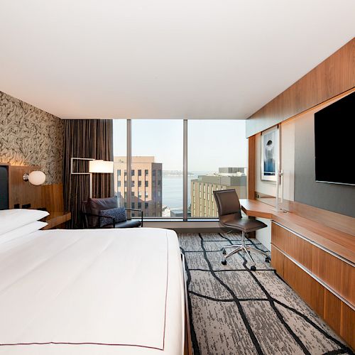 A modern hotel room with a large bed, flat-screen TV, desk, chair, and a view of city buildings and water through floor-to-ceiling windows.