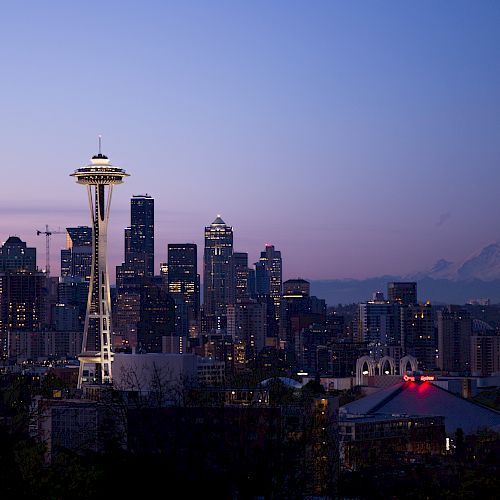The image shows the Seattle skyline at dusk, featuring the Space Needle, downtown buildings, and Mount Rainier in the background, under a clear sky.