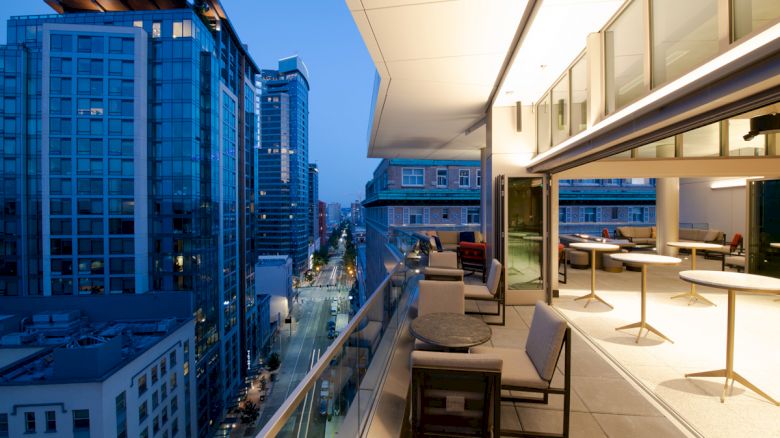 A modern balcony view at dusk, overlooking a city street with tall buildings, featuring outdoor seating and tables on the balcony.
